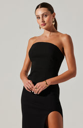 A woman is wearing a strapless black dress that fits snugly and has a thigh-high slit on the left side. She accessorizes with gold hoop earrings, a gold bracelet on her right wrist, and several rings on her fingers.